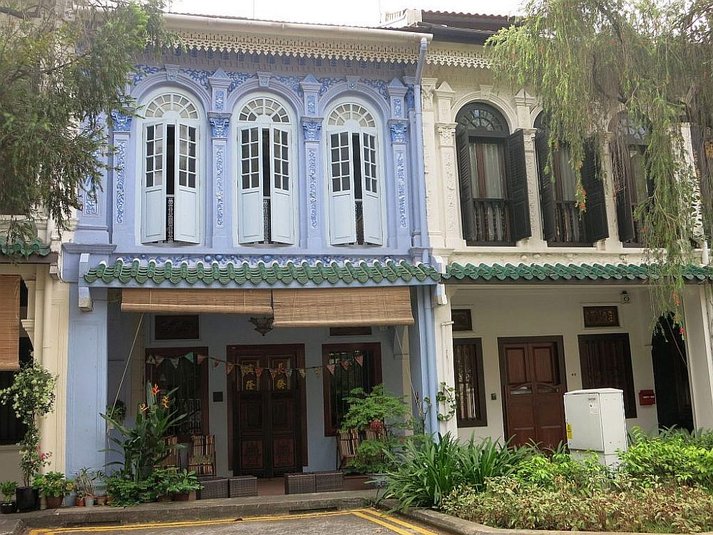 Two row houses in Singapore: each is two stories high, with the ground floor shaded by a small roof. The upper floors are quite decorative, with tall windows with shutters and arched windows at their tops, and ornate decorative elements between and above the windows. The left-hand house is predominantly a pastel purplish-blue and the right-hand house is beige with brown edging and shutters. I think Singapore would be a great place to live as an expat.