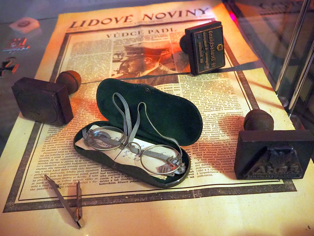 The newspaper underneath is in Czech, titled "Lidove Noviny". The article's title is "Dudce Padl" and the only photo of the page is of Hitler. On top of the paper is an open glasses case with small round wire-rimmed glasses, three stamps and a tie pin.