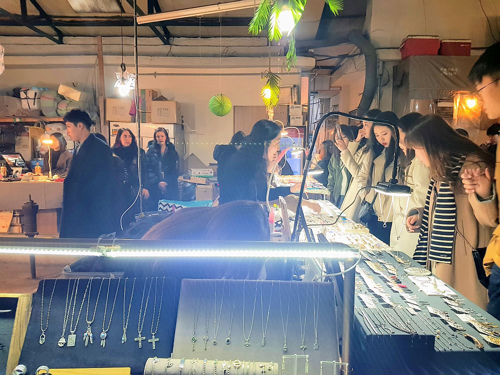 In front is a lit case in which necklaces are displayed. Along the right a group of people, mostly young women, are lined up looking at small items (jewelry, I think) on display on tables. On the left part of another stall is visible with a few people walking by.