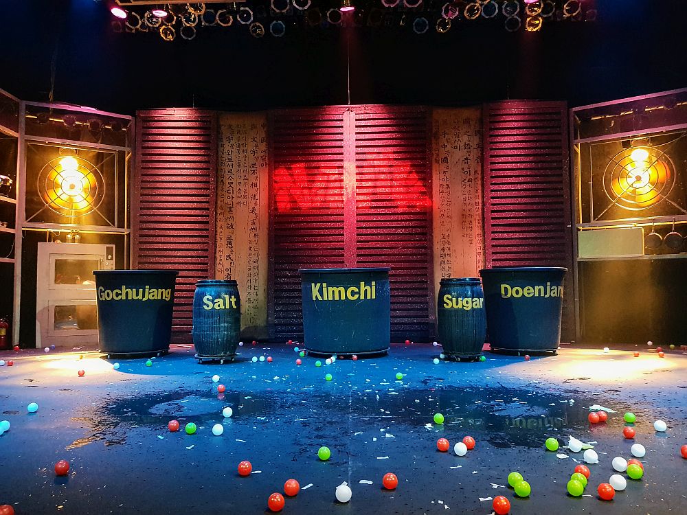 The stage of the Hongdae Nanta Theatre in the Hongik University Street neighborhood. There are no people on the stage, but the word Nanta is spraypainted on the back wall. In front of the wall are 4 big barrels of various sizes, each labeled: GochuJang, Salt, Kimchi, Sugar and Doenjan. Between them and the camer are scattered small balls: red, white and green, on a black wet floor that looks like it's wet in spots.