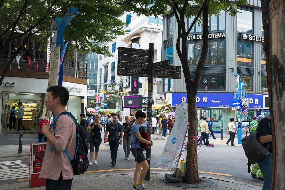 A view of an intersection of Hongik University Street. People are walking both in the street and on the sidewalks in all directions. Many look like students, carrying backpacks. Trees on the corner in the foreground, buildings in the background with shops on the ground floor. 