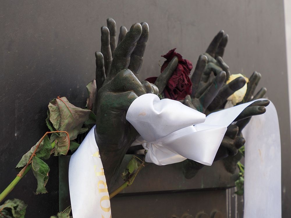 A metal plaque on a plain background. The cluster of hands emerges from the top of the plaque: about 7 hands, some showing the V sign with palm outward, some showing all fingers with palm outward. A white ribbon has been wrapped around some of the hands and obscures them, along with a shrivelled rose. It emphasizes the peaceful nature of the protests that started the Velvet Revolution against the communist leadership in Prague.