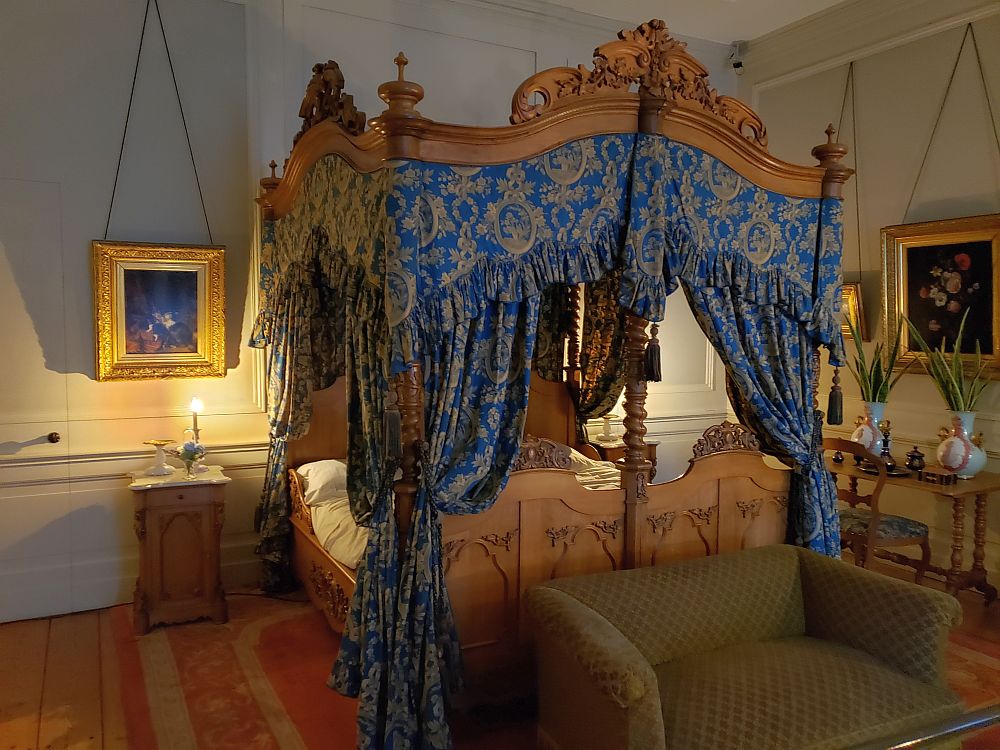 The bed is a four-poster bed made of wood, all decoratively carved. It has rich fabrics hanging down in blue with a brown design. Paintings of flowers hang on either side in gold frames and a rather simple brown sofa stands at the food of the bed. Besides the paintings, the walls are quite plain, painted rather than papered.