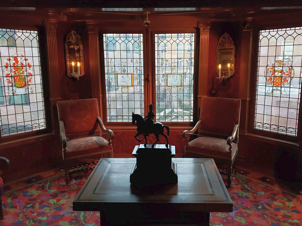 A square dark wood table stands in the center of the room, with a small pedestal in its midde, holding a statue of a man on horseback. The floor has a dark red, flower-patterned carpet. Beyond the table are two simple dark red armchairs. Between them, two paned windows and one window on either side of them as well. The four visible windows have small panes that have been painted. The paintings in the middle two windows (in a picture at the beginning of this article describes one of them) are hard to see, but the two one either side are painted brightly: on the left, a vase with bright orange flowers. On the right, a coat of arms, also in orange.