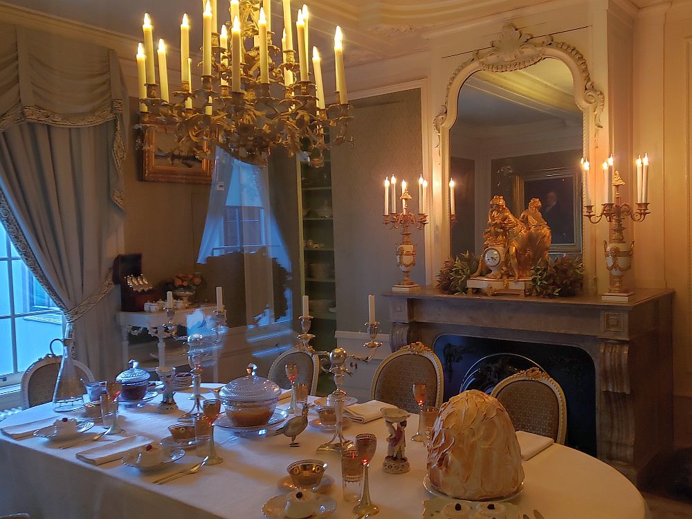 The table is set with a white tablecloth and napkin, silverware and fine china. The chandelier is smaller and simpler than in the ballroom. The mantelpiece is also simpler, with a baroque-looking gold clock sitting on it, along with two candlesticks, each holding 5-6 candles. A large mirror backs the clock. 