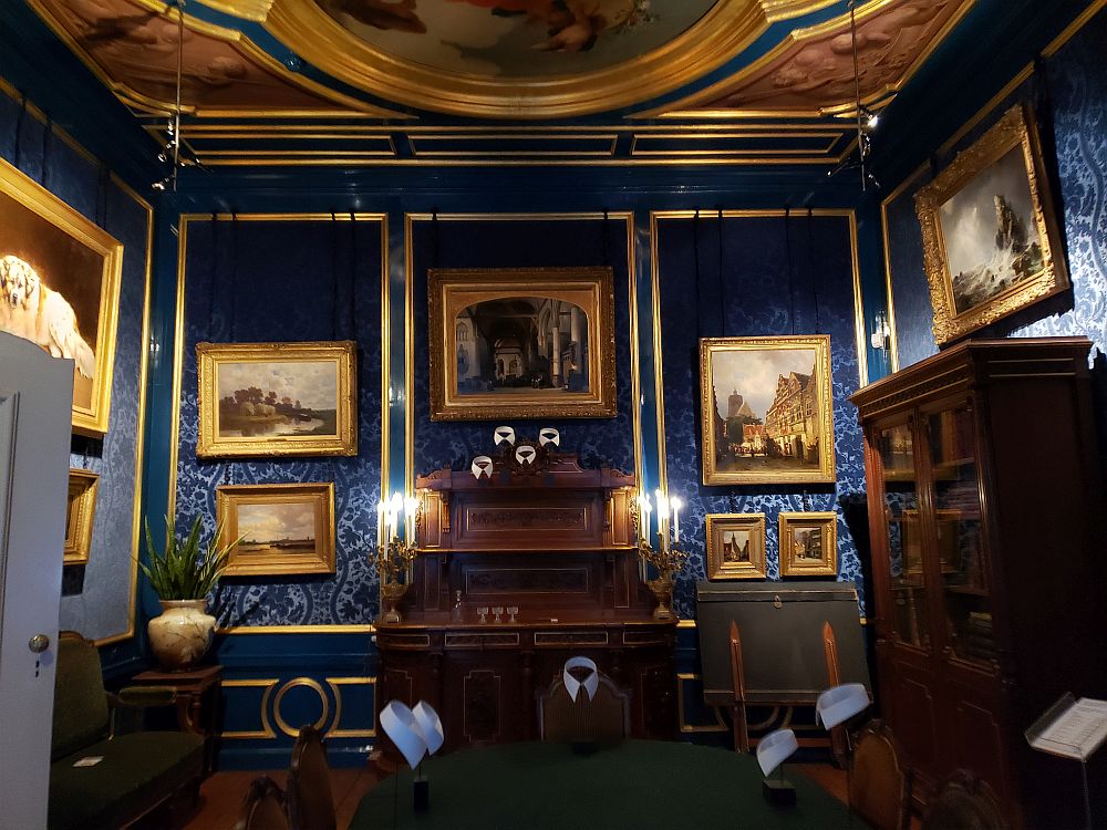 The walls of the parlor are covered with a royal-blue velvet paper. The edging is gold, as is the ceiling. A small part of the central painted ceiling is visible. On the wall panels is a collection of Golden Age paintings, each framed in gold. 