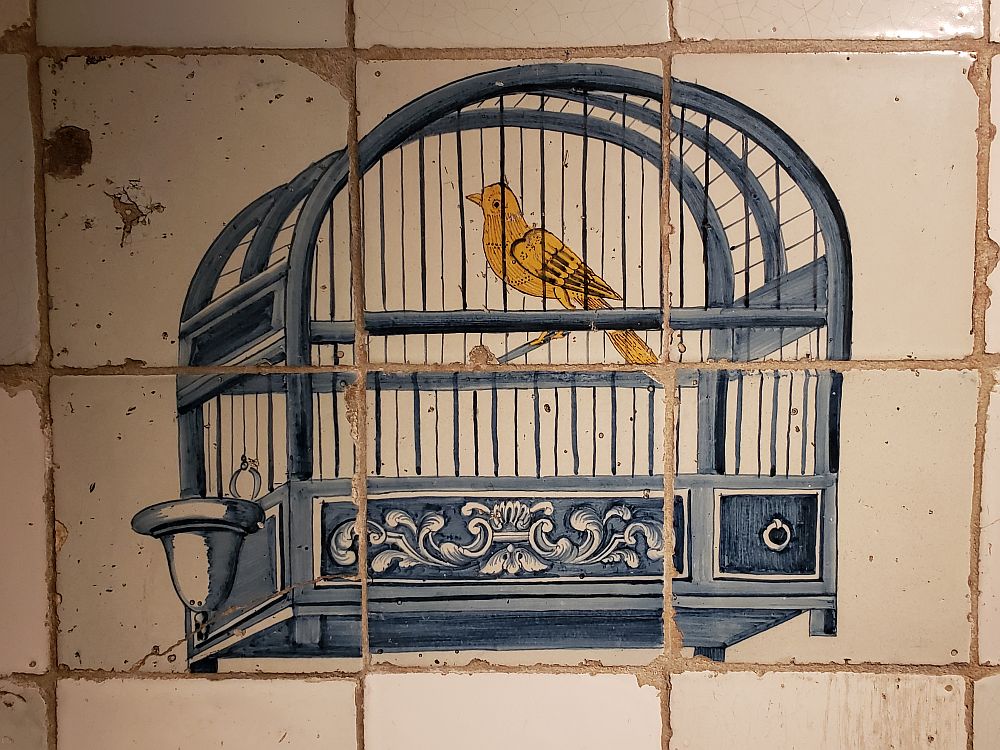 Covering a space of six tiles, the image is of a blue bird cage, quite simple, holding a single yellow bird. Other than the image, the tiles are white and a bit chipped around the edges.