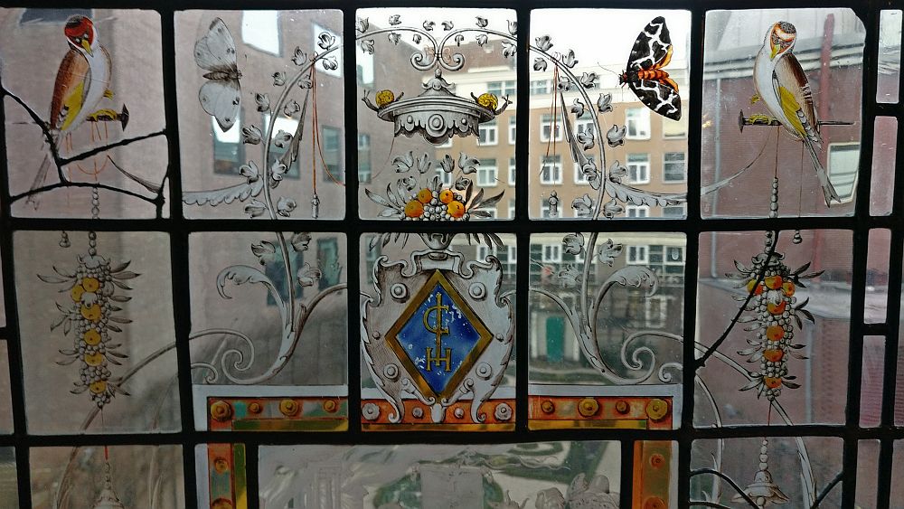 The glass is divided in rectagular panes. The center one has a small coat of arms with a brown frame and blue background. It looks like a decorative form of C, I and H on the blue background. Sitting on top of the coat of arms is a vase with orange flowers, and above that, an odd little silver roof, with a snail on either side. Other animals are on nearby panes: a bird on either side, a butterfly on each side too. Also a bunch of what looks like grapes, but with orange flowers hangs on either side of the coat of arms. Between the paintings, the glass is clear, allowing a view of the garden. 