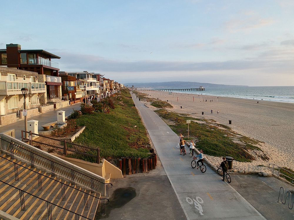 On the left, a row of houses, modern and closely set together, extending into the distance. In the center, the paved bike path with a few bikers on it: it goes straight into the distance parallel to the row of homes. Between the home and the path is a strip of low scrub-covered land, with a stairway crossing it in the foreground. On the left, also extending into the distance is the beach, with a bit of the sea visible in the distance. 