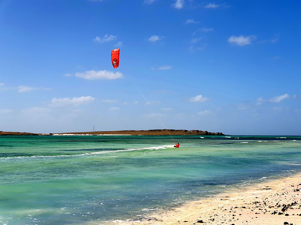 A man is leaning (perhaps falling) in the shallow water near a beach. His kite nearly above him is bright red.