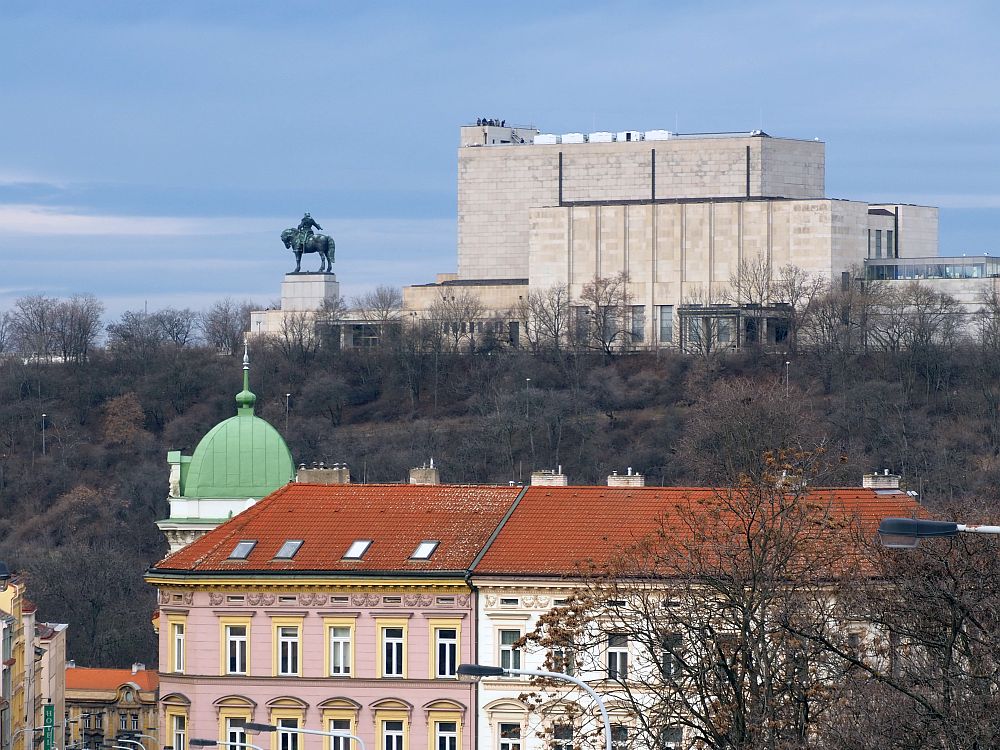 A very large block of a building on a rise, with an equestrian statue to its left. The statue must be quite large to be so visible from such a distance. Below the ridge are some more typical older buildings: one in pink with yellow edging and one in white, both with decorative elements around the window. A green dome peeks from behind one of the buildings. The side of the hill between the older buildings and the mausoleum is tree-covered.