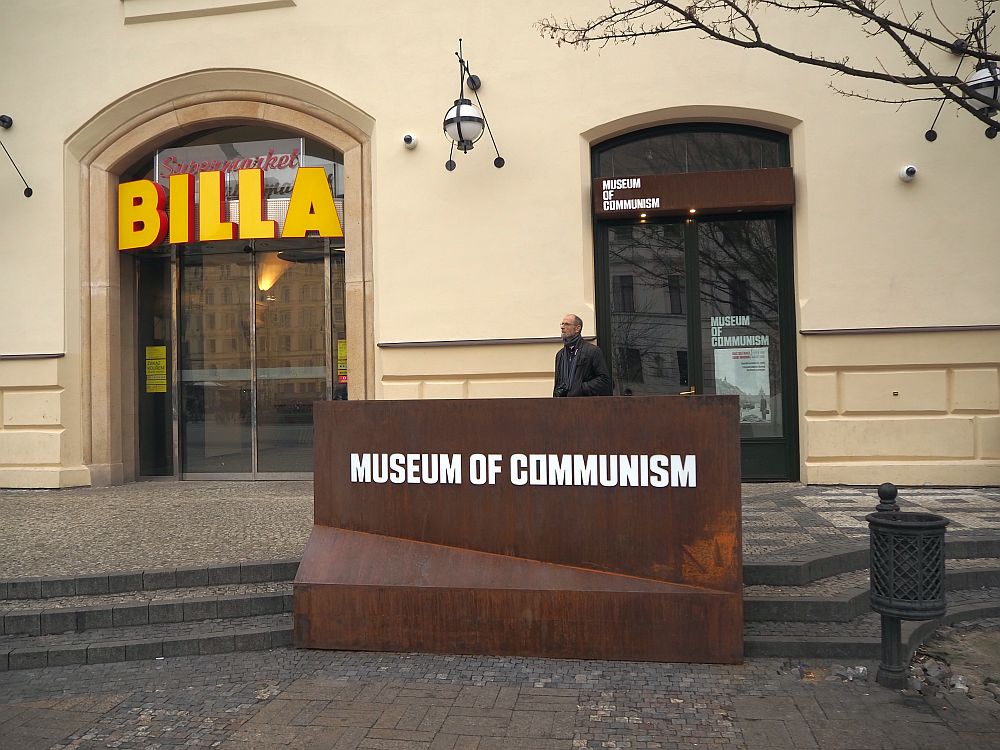 A plain metal sign reads "Museum of Communism". Behind that is the simple entrance to the museum. To the left of the entrance is a bigger arched door, with big letters proclaiming "BILLA": the entrance to a supermarket. One man, my husband, stands in front of the museum entrance, waiting for me to finish taking pictures.