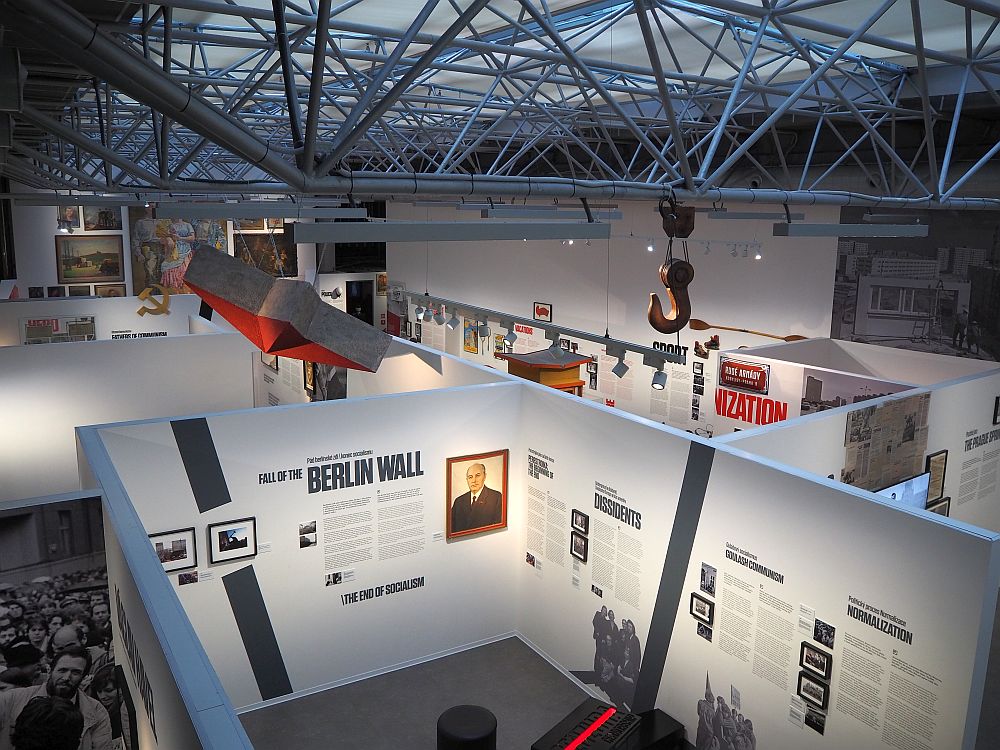 The space has a high roof with a latticework of bars holding up a translucent glass ceiling. Below, the walls around the exhibit spaces are normal wall height but without ceilings, so from this viewpoint the contents of the rooms are visible. The nearest room has titles painted on the wall that read "Fall of the Berlin Wall", "Dissidents" and "Normalization." The rest is not legible form this distance.