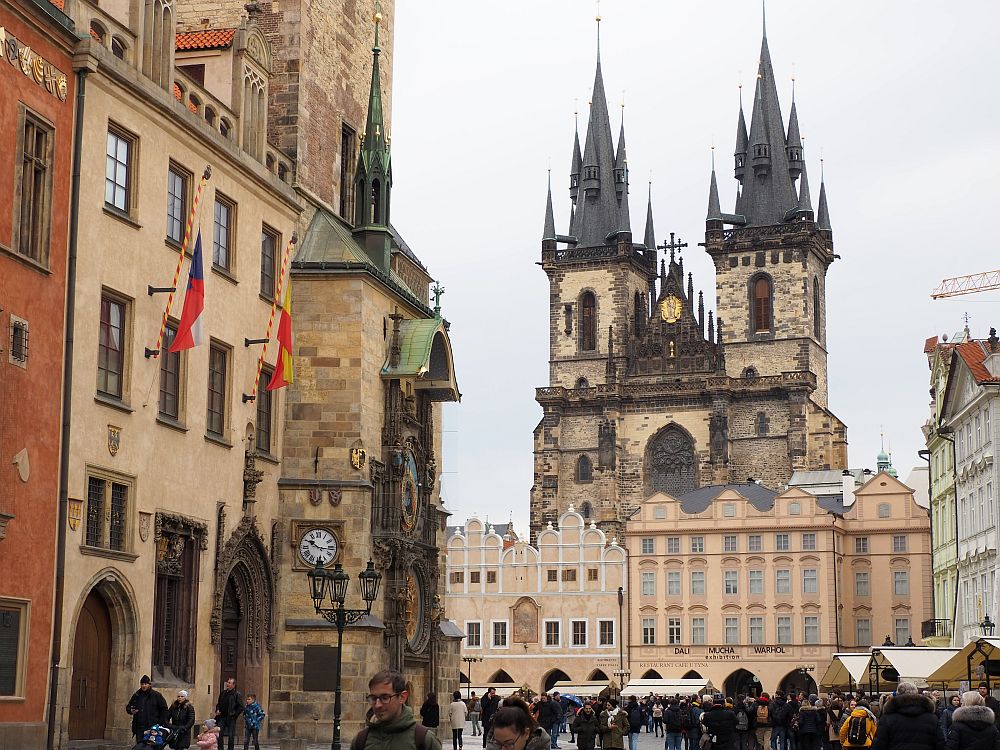 On the left is the famous astronomical clock, built onto the side of a building. In the background, across a big open square, is a row of buildings, about 6 stories high, all in shades of beige. Behind them and much taller: a large cathedral with two spires, each adorned with a number of smaller turrets. At least the communist rulers of Prague left the old section intact.