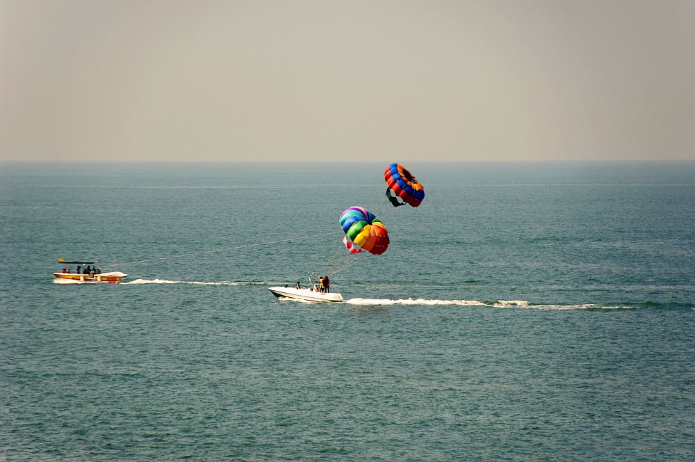 In this phot are two boats. The one in the center has two parasails in the air above it, or perhaps the boat to the left is pulling one of them. It's hard to tell. Both boats appear to be moving, but not very fast, and the parasails are multi-colored.