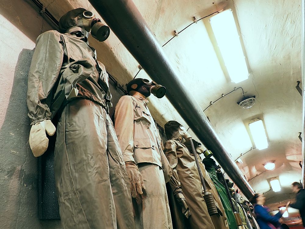 Taken from floor level, looking at a row of mannequins, each dressed in what looks like rain jackets and pants and wearing a gas mask and mittens.