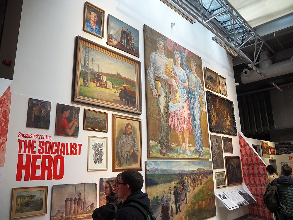 The wall is about two stories high and is covered with artworks, mostly showing things like proud peasants and workers in idealized country settings. A few people at the bottom of the photo look at them. The title painted on the wall reads "The Socialist Hero"