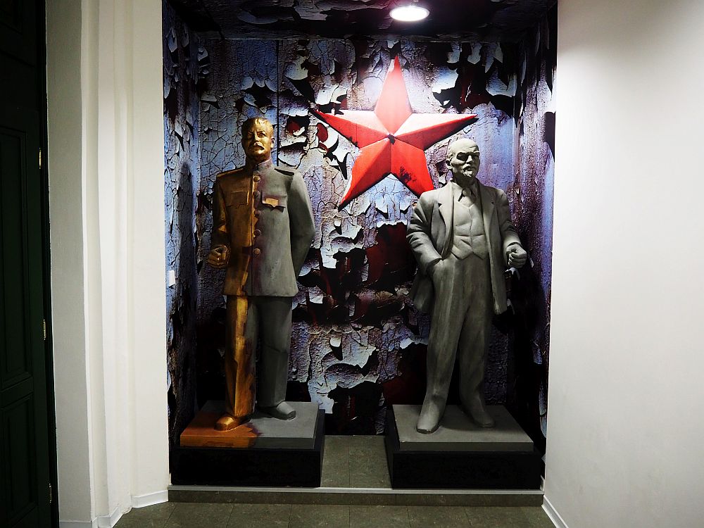 Two full-size statued in a niche in the wall: Stalin on the left in a military suit, Lenin on the right in a civilian suit. Both seem to be made of metal. The wall behind them has badly peeling paint and, in the middle, between the two statues, is a big red star.