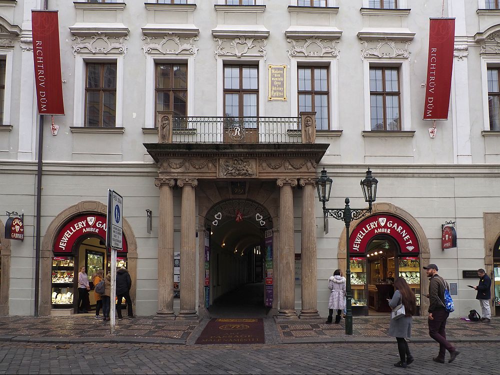 A typical Prague building with ornate touches above the windows. In the center ground floor is a grand entrance with two pillars on either side supporting a balcony. On either side of the entrance are shops, both with a sign reading "Jewellery Garnet"