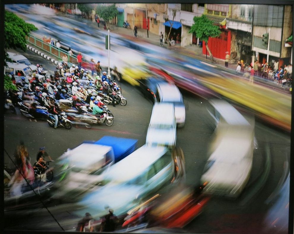 A cluster of scoooters stand still in the middle left. Top left to middle right and top  left, curving around to bottom left: cars stream by, all blurry from motion. In the background: the shops along the street are also still. 