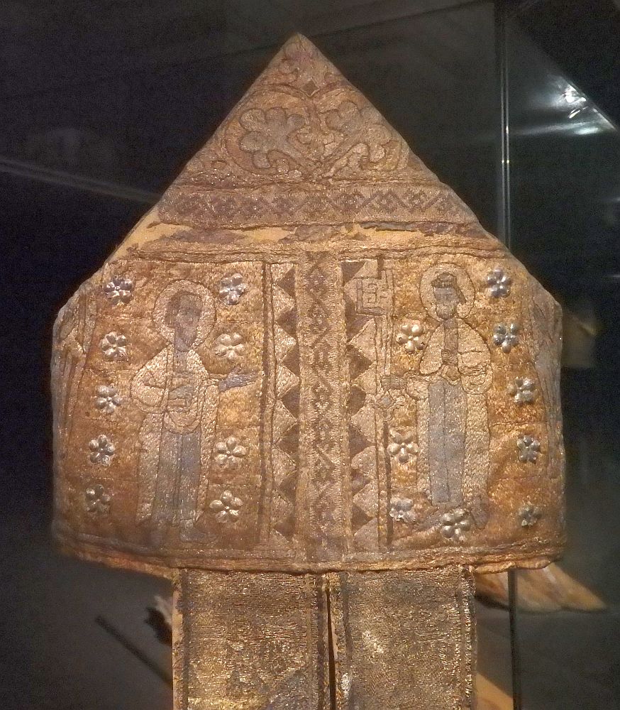 The mitre is square with a triangular point on top and two wide ribbons hanging down. It is entirely covered with embroidery, mostly flowers and abstract shapes except for two men, one on each side, each with a halo.