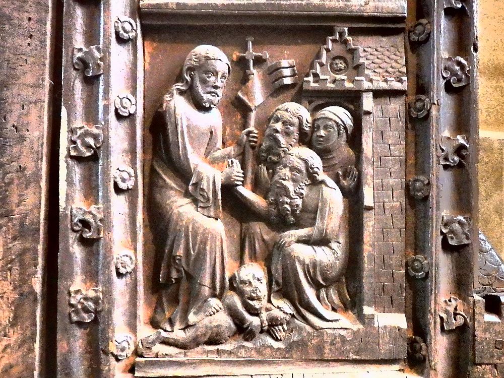 A bas-relief depicts a man on the left, standing, holding a staff with a cross on its top, with one foot on a chained creature - a dog? a devil? It's not clear. Two men and a woman huddle on the right, and the man on the left holds the hand of one of the men.