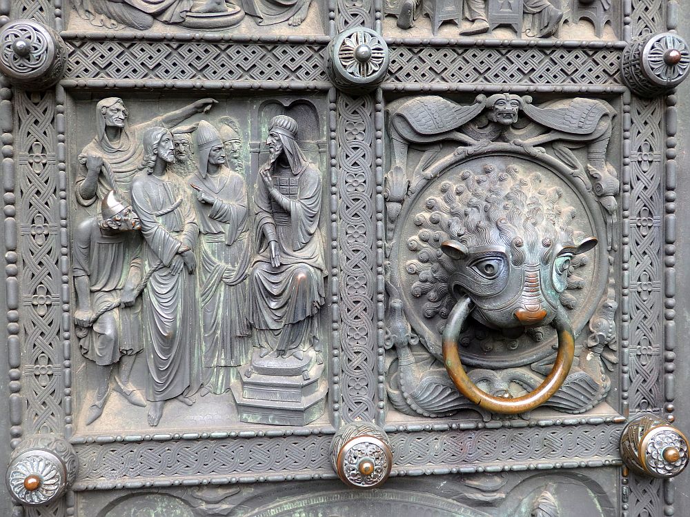 Detail of the metal door. The left panel is a bas-relief showing a person (Jesus?) sitting on a throne, speaking to a group of men. The right panel is the door knocker: a creature, maybe a lion, with the ring of the knocker in its mouth.