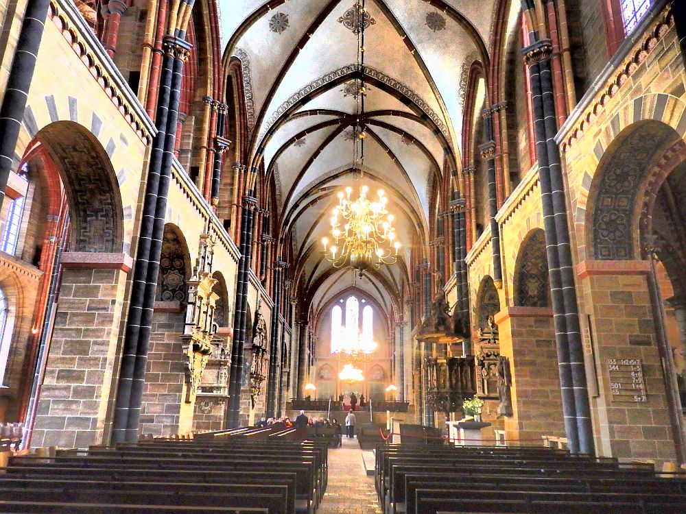 A typical medieval cathedral structure, with columns along the sides and gothic arches in the ceiling. Photo looks the length of the nave from the center aisle between the pews, to the altar in the far distance, blurry because of the light coming in the windows behind the altar.