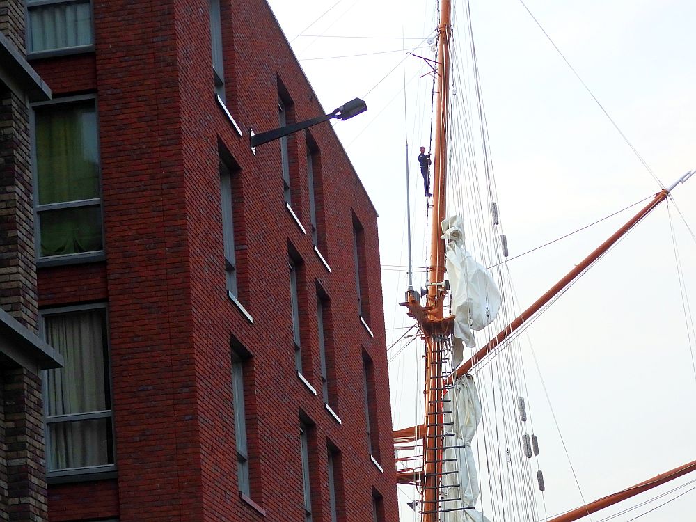 Looking past a modern red-brick building (offices, probably), a mast of a ship is visible (not a tall ship, I don't think). A man stands more than half-way up the rigging. It looks like he's just standing on a bit of rope. 