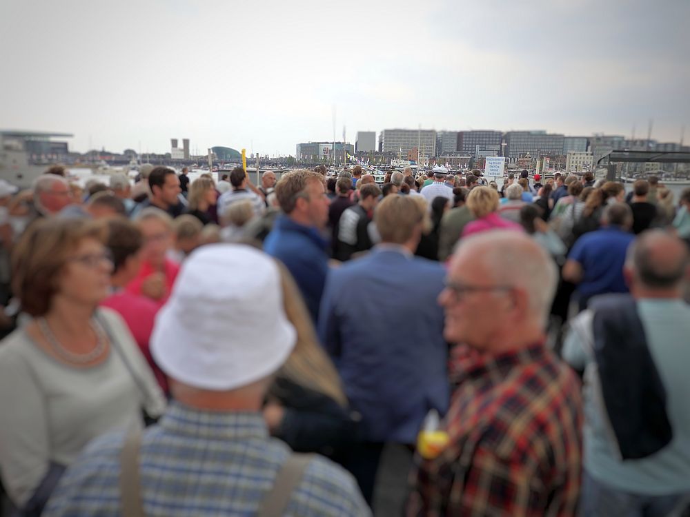 A crowd of people all standing and waiting. The dock is not visible in the background because of the crowd, but a group of apartment buildings are visible in the background.