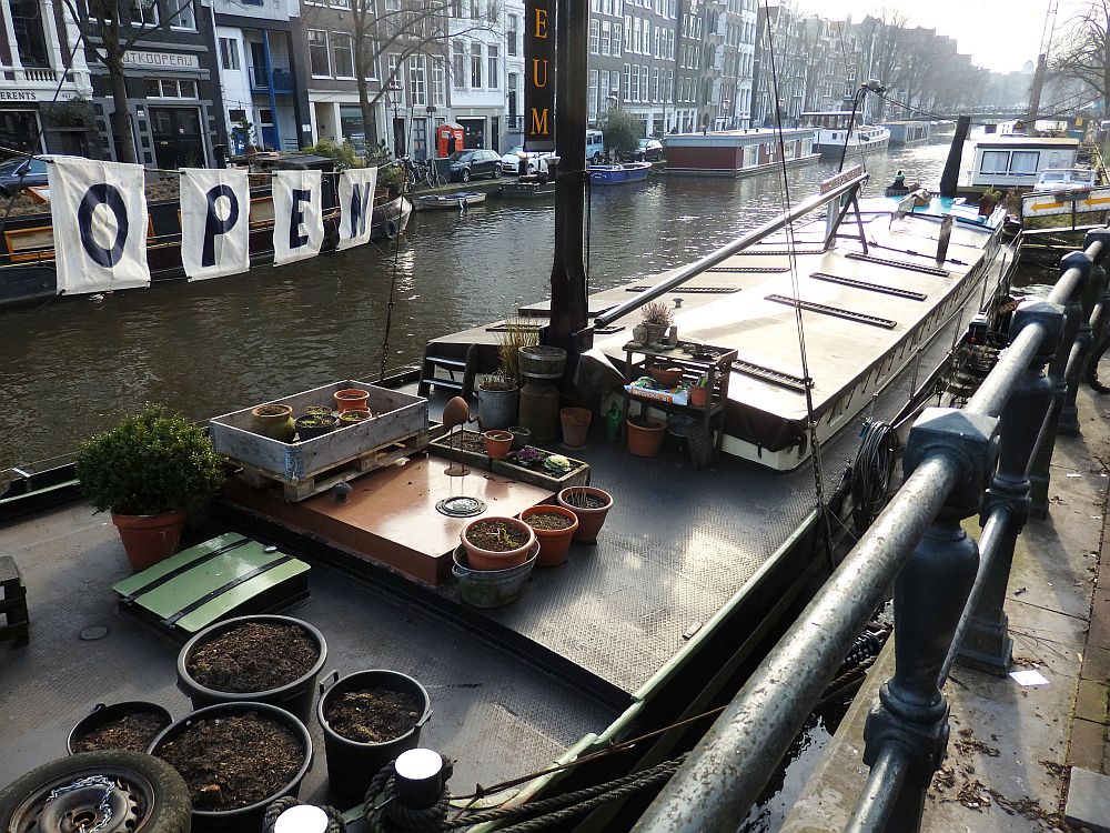 Exterior view of the houseboat. It's low to the water and very long and narrow. On the roof are a bunch of flowerpots; only a few with plants since the photo was taken in the winter. Beyond the boat is a view down the canal, with some other houseboats visible along both banks.