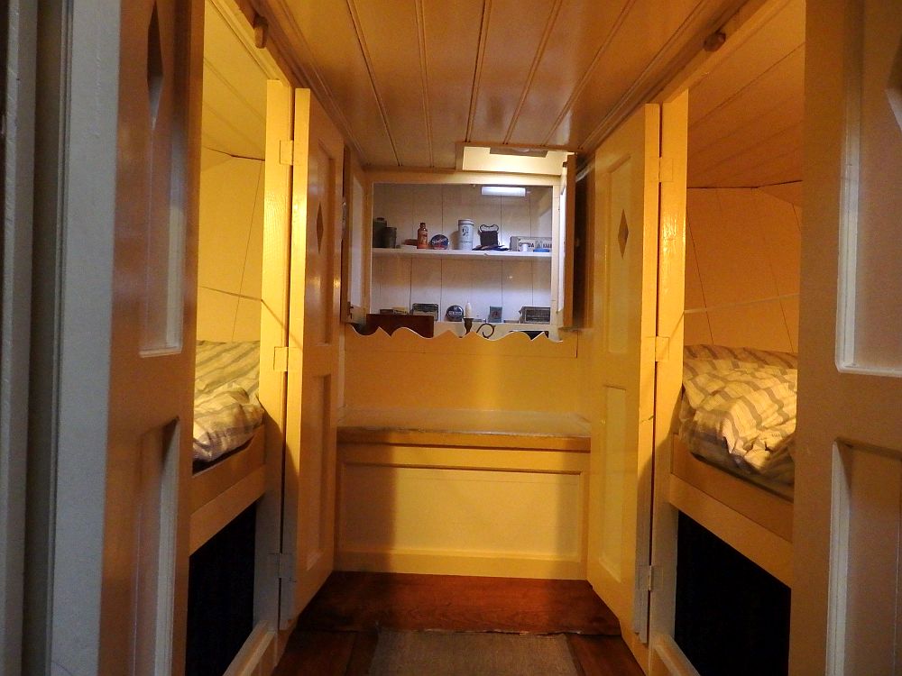 A hallway straight ahead that ends in a wall where a small cabinet with glass front hangs. Two shelves inside have various small objects. On the left and the right are two openings, each with a bit of the bed visible: a mattress on a shelf, parallel to the hallway.