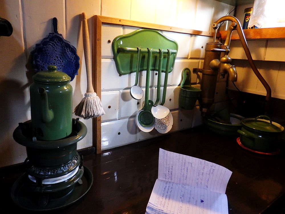 A round stove on the left has a green coffepot on it. A green metal rack has four utensils (ladles) hanging from it. In the corner is a waterpump: the kind you have to pump a handle up and down to get the water up. 
