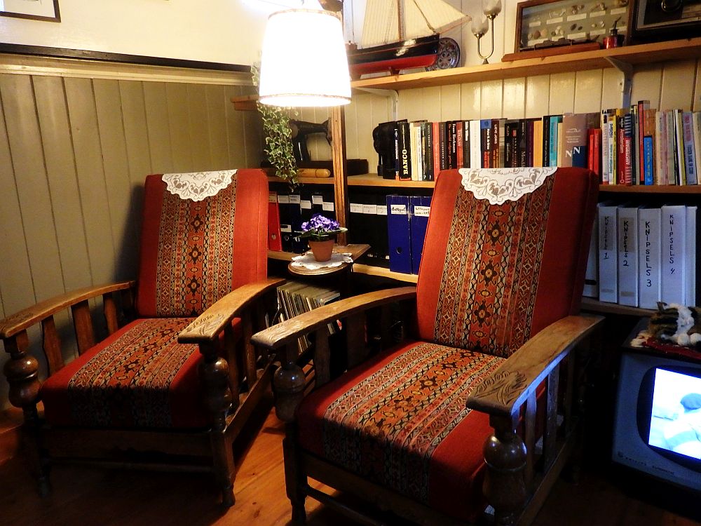 Two wooden chairs with thick red cushions for seat and back, and doilies on the tops. Behind them is a bookshelf lined with books, and, on the top shelf, a model sailboat.