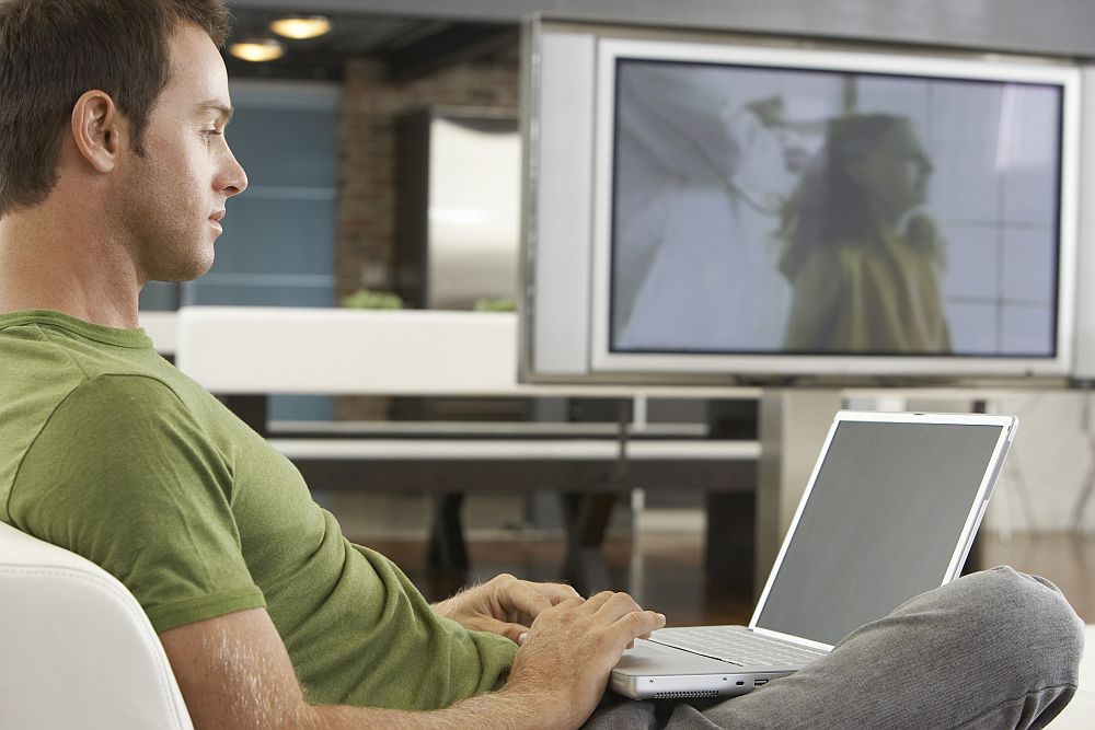 A white man wearing a green t-shirt sits sideways to the camera on an armchair or sofa. He looks at a laptop in his lap. In the background, a large tv is on, showing a vague image of a woman. 