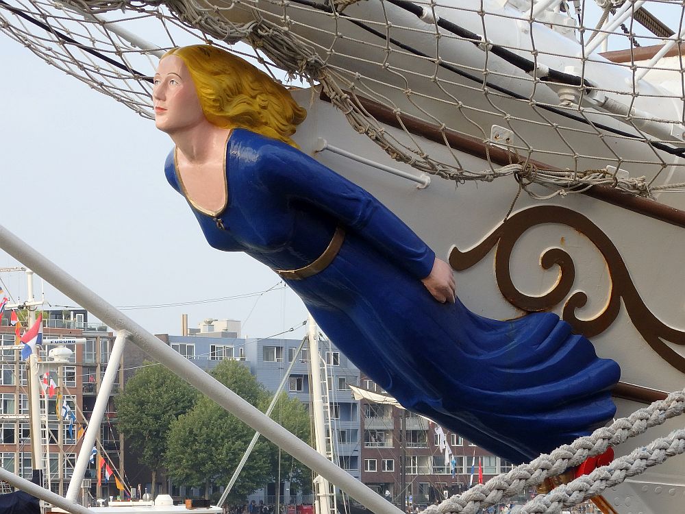 A figurehead of carved wood in the shape of a woman. She is carved under the bow of a ship, so her back is attached while her front is visible. She wears a simple long, blue dress with a scoop neck. She is white with blond hair streaming behind her. 