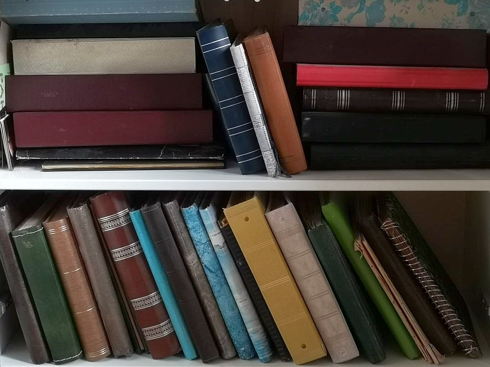 two rows of photo albums of various shapes, sizes and colors. On the top shelf they are mostly in two piles, with a few smaller ones standing between the piles. On the lower shelf they are all upright but leaning to the left. 