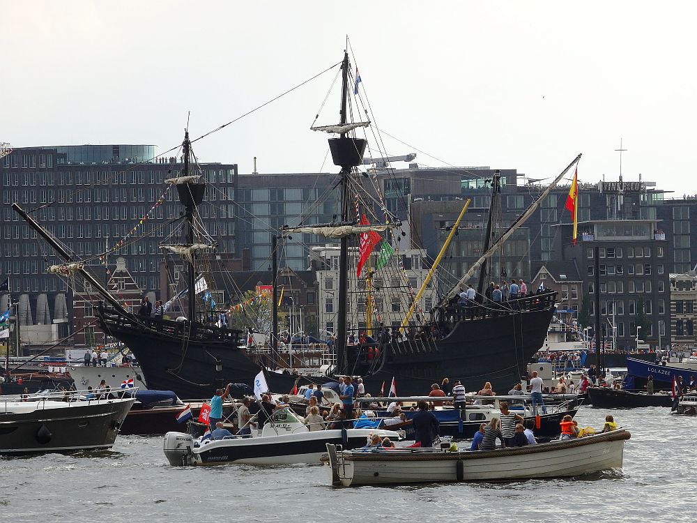 Another general view. In the background, a row of modern apartment buildings. Moorred on the other side: another ship that looks like a pirate ship: black with a high bow and high stern, on which people are standing. It has two masts, each with a "crow's nest" on the top. In front of that, on the river, lots of small motorboats pass, crowded with passengers.