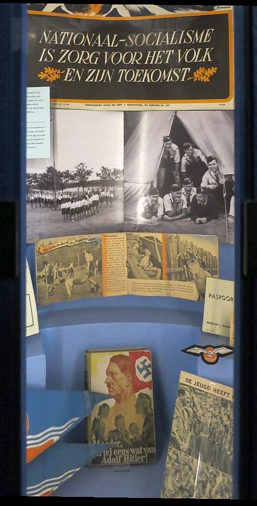 The display is tall and narrow. At the top is the slogan translated in the caption. Under that, photos of Nazi youth lined up in a field on the left and a group of young men in informal uniforms looking out of a tent and smiling on the right. Below that, some publication open to a page of photos of Nazi youth doing activities. On the floor of the display stand several books.