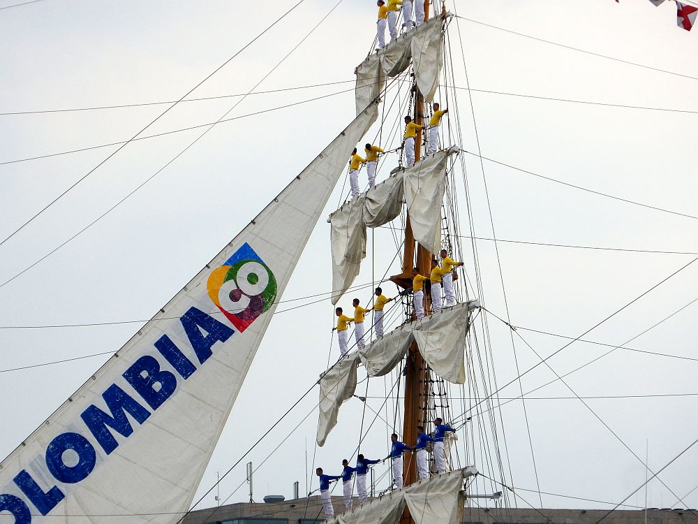 A close up of a mast of a tall ship, marked as Colombia on the one sail that is open. Along each horizontal strut, standing on the furled sail hanging from each strut, is a line of men. The top three rows (6, 4 and 4 people) wear white pants and yellow shirts. The bottom row (6 people) wears white pants and yellow shirt. 