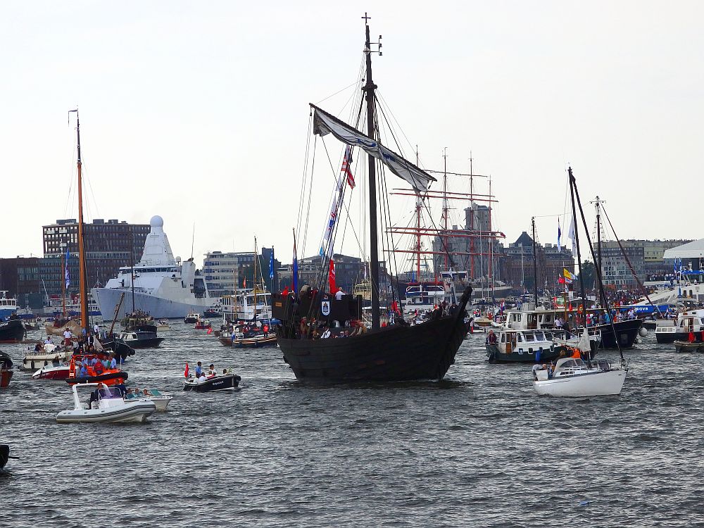 Lots of ships and boats in this photo. The biggeset looks like a small black pirate ship with a high bow and stern and a single mast with a horizontal sail that is furled up. Around it are lots of smaller boats: motor boats and sailboats of all sizes. 