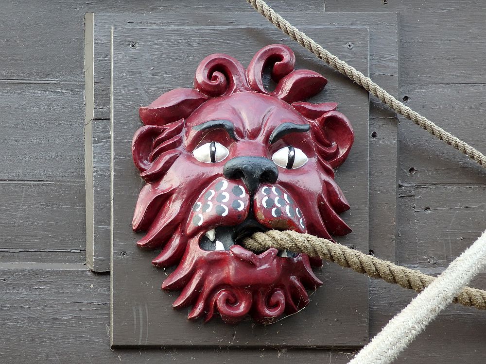 Closeup of a wooden carving, painted red, of a lion's head. It has a hole in its mouth for a rope to go through. 