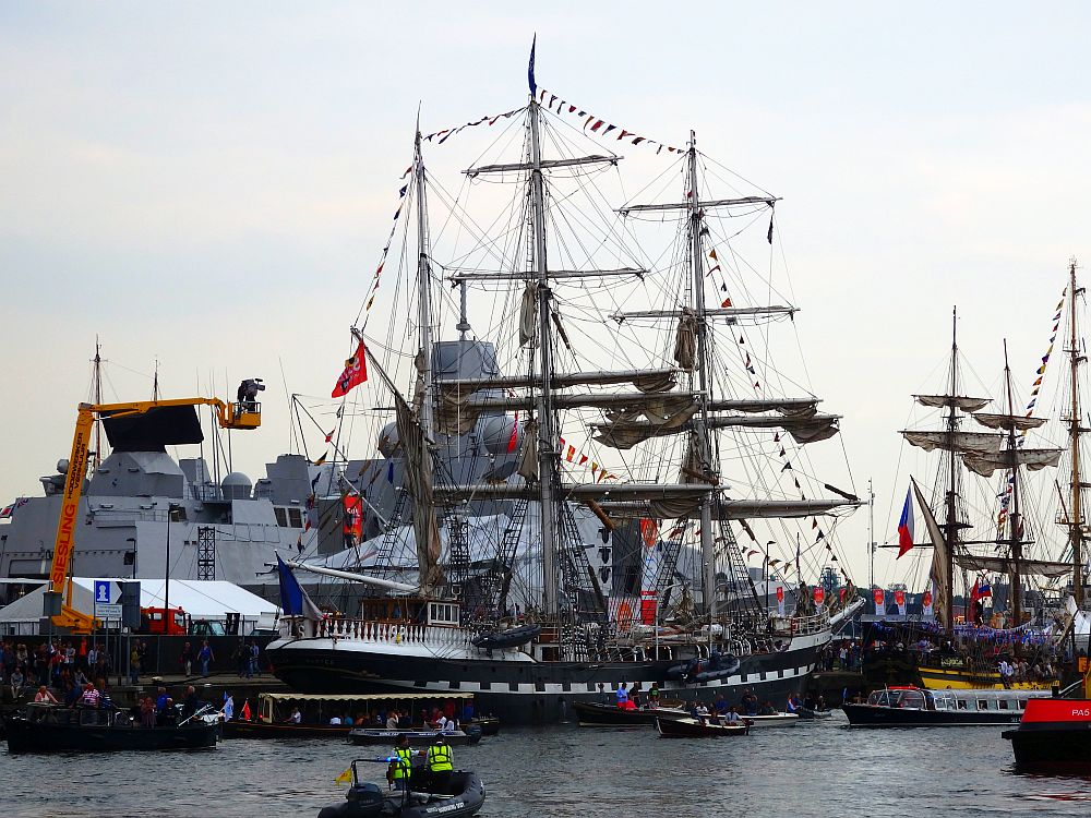 A tall ship with three masts, with small decorative flags strung along the top of the three masts and conneting to the bow and stern of the ship. Behind it is what looks like a military ship, modern and angular and painted grey. 