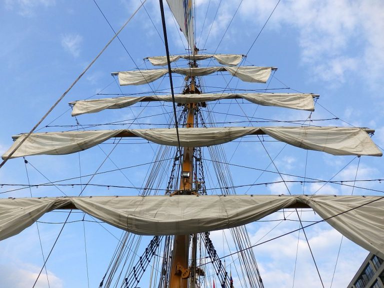 Looking up at a tall ship mast with its sails furled on horizontal spars.