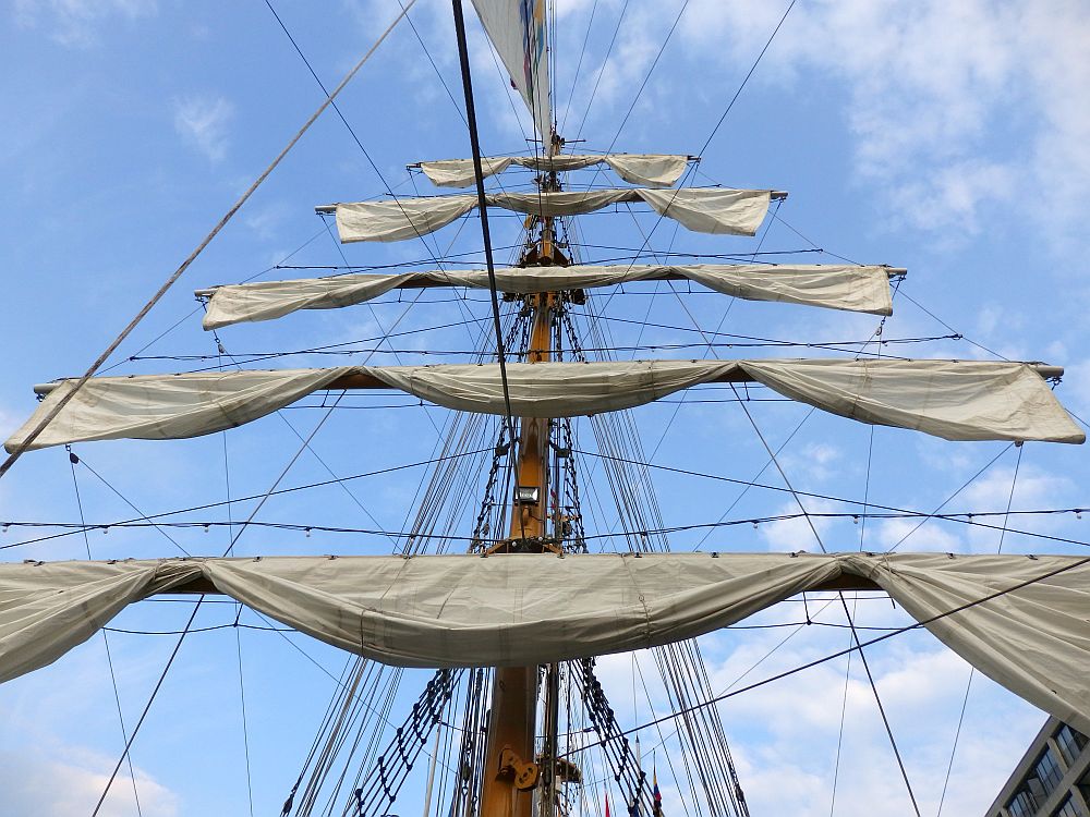 Looking up at a tall ship mast with five sails, furled on horizontal beams, one above the other.