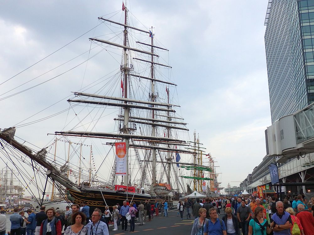 Facing down a long straight wharf, many tall ships are moored along its side, so this creates a row of masts into the distance. The wharf is full of people walking, and a bit of one building is visible to the right of the wharf.