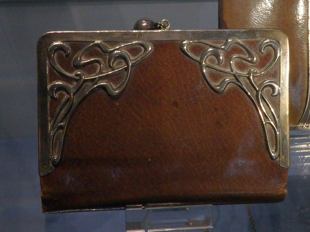 A smooth brown leather bag with a metal rim on the top and sides. The metal has art nouveau-style swirls in the top corners.