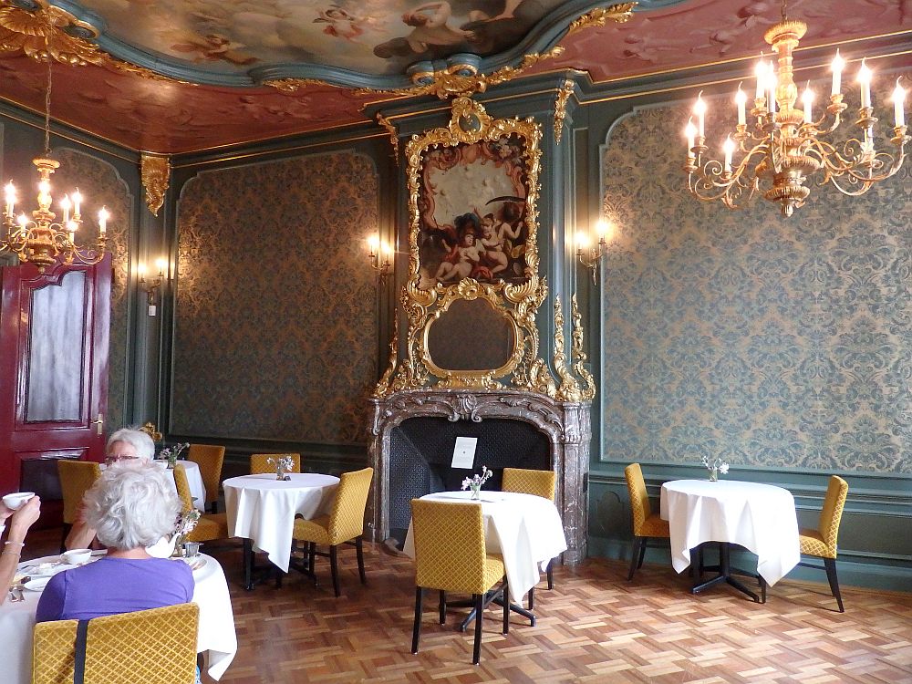 The room has a high ceiling, and the bit that is visible has a gilded inset frame with a baroque painting in it. The mantelpiece is also gilt-edged, and the gold extends around an oval mirror and a baroque painting above the mirror, with a gilt-framed crest of some sort above that. The walls are comparatively simple, with a blue-flowered wallpaper and a wainscoting below that. the floor is parquet and a few tables with tablecloths are scattered around. Three women sit at a table on the left.