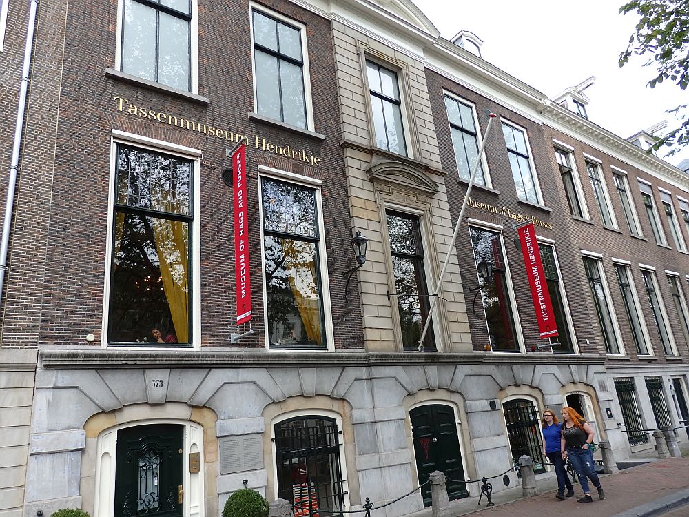 The front of the museum is quite grand-looking: red brick with tall windows (smaller on the ground floor). Red banners extend from the facade advertising the museum. Two signs say "Tassenmuseum Hendrikje" and "Museum of Bags and Purses"