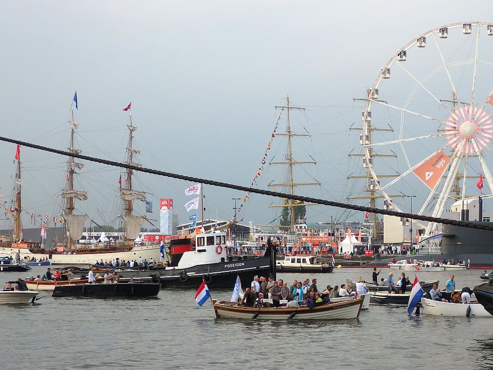 Another general view of the IJ river: Lots of boats passing to the left or the right. In the background a couple of tall ships are visible, moored on the other side. The boats on the river are mostly small, open motorboats. One looks like a tugboat or fishing boat. In the background, part of a ferris wheel is visible.